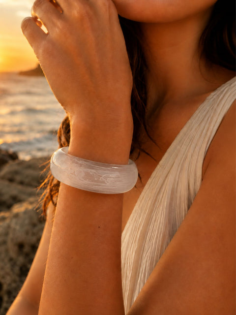 Zeni wide acrylic bangle in white marble displayed on natural coastal rocks at sunset