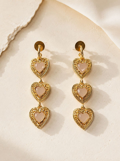 A pair of gold drop earrings with three stacked heart-shaped charms, each featuring a pale pink stone in the center.