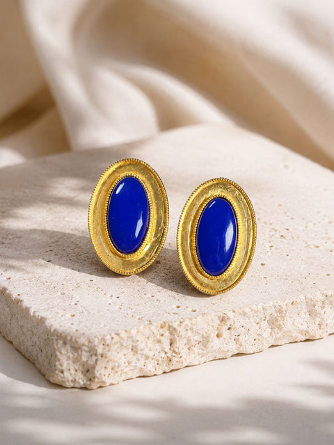 Gold oval stud earrings with glossy cobalt blue stones displayed on a natural stone.